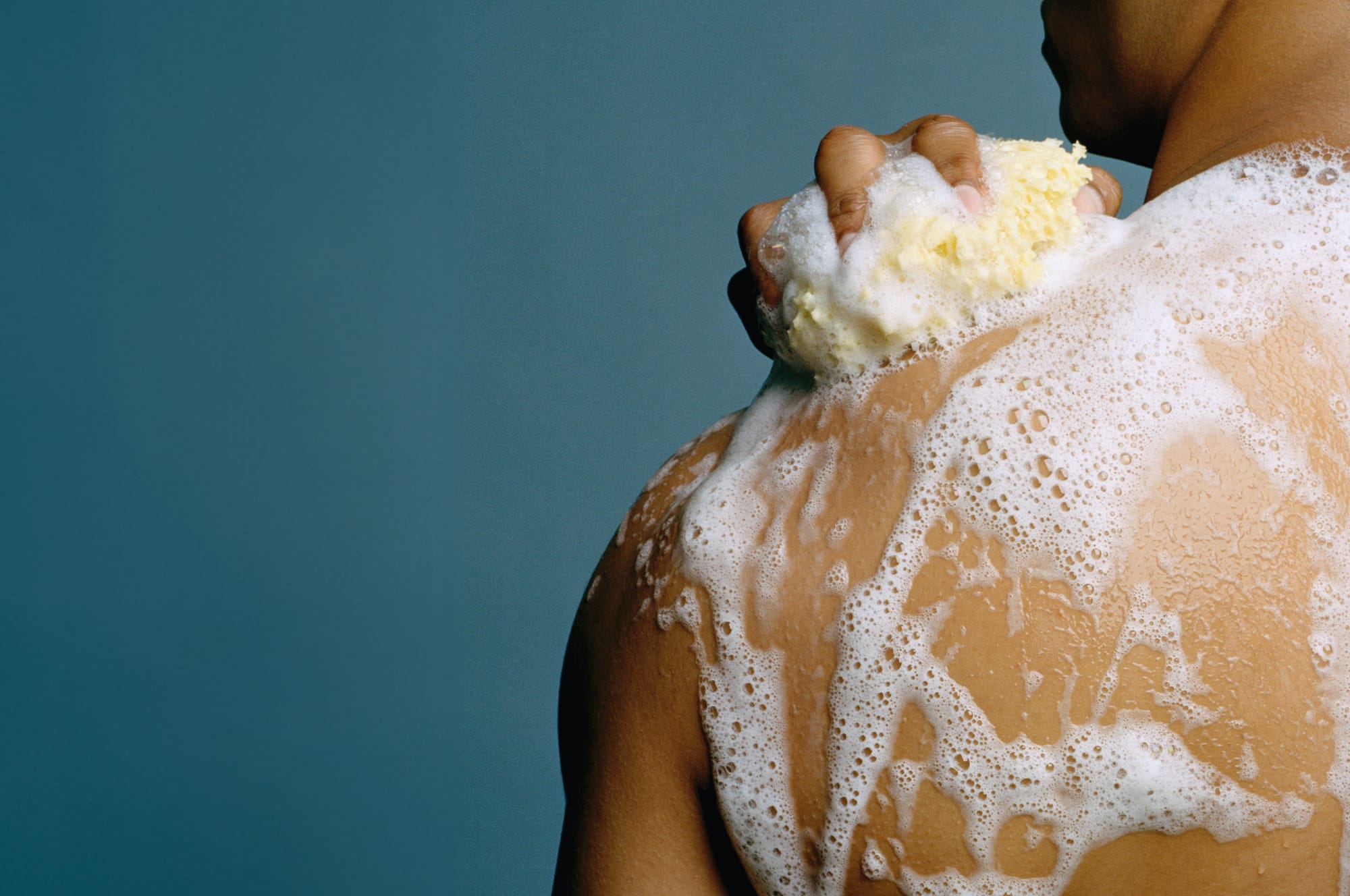 Young man using a sea sponge to soap his body while showering.