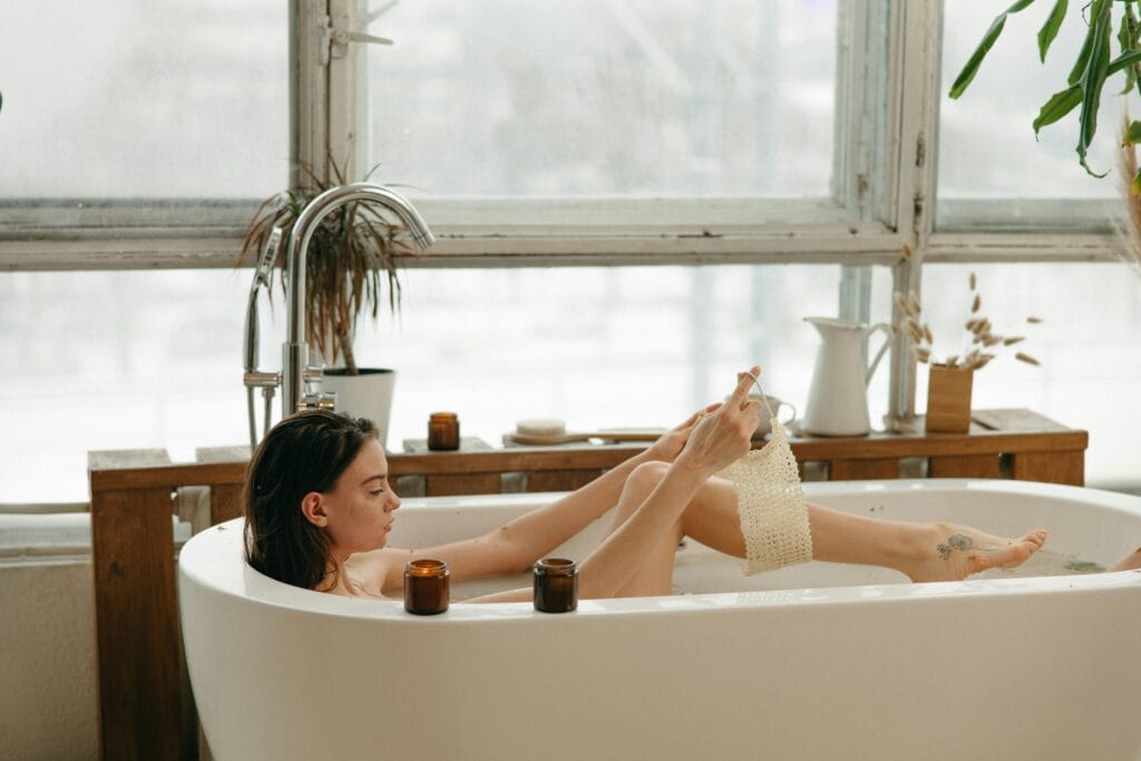 7 Bath Essentials to keep in your Bath Time Woman in a bathtub with candles, relaxing and using a body scrub. Cozy indoor setting.